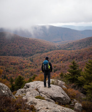 Backpacker male hiker standing on mountain rock peak enjoying view of misty colorful forest valley in autumn