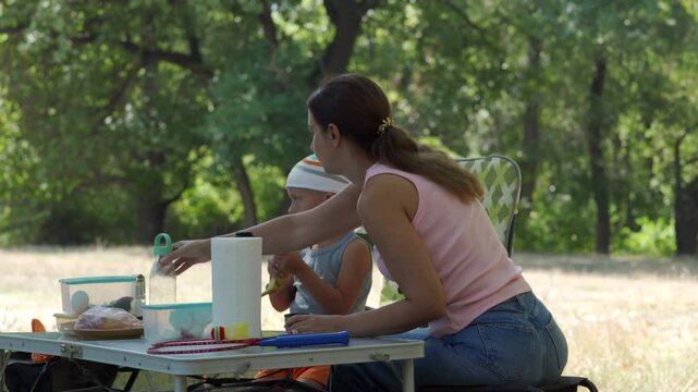 A peaceful mother watches her focused son peel a banana at a picnic table. They share a quiet, loving moment together. A beautiful green park surrounds them.