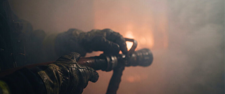 Hands gripping firefighting equipment in a smoky emergency environment with soft firelight in the distance. Suitable for commercial safety materials.