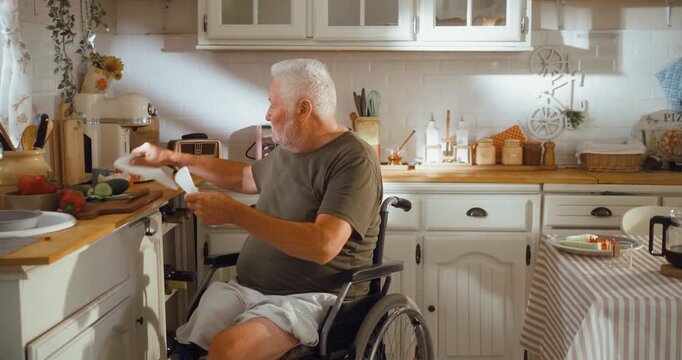 Senior man in a wheelchair wipes his hands with a napkin in the kitchen before breakfast, showing an independent and positive morning routine at home.