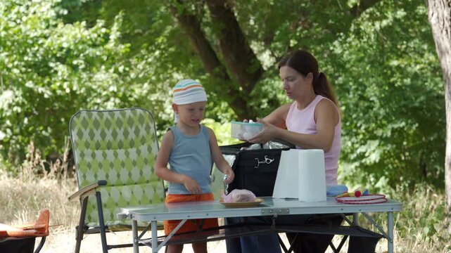 Woman arranging picnic food and utensils at portable table while boy in striped hat stands nearby with plate, creating calm family moment in shaded park clearing with green trees and casual leisure