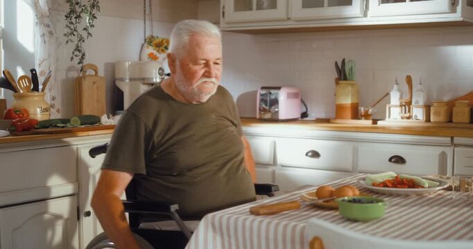 Senior man in a wheelchair approaches the kitchen table and begins breakfast by peeling a boiled egg, showing independent daily life at home.