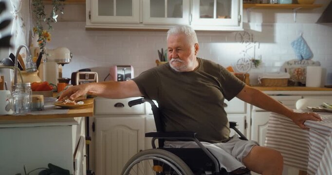 Independent senior man in a wheelchair clears the kitchen table after breakfast, showing active daily life and self sufficient living at home.