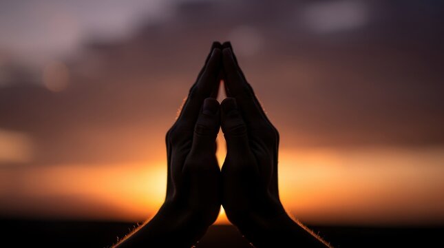 Silhouetted hands pressed together in a prayer or meditation gesture against a warm, glowing sunset sky.