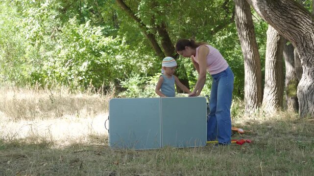 A cooperative mother and her helpful son fold up a camping table together. They share a peaceful, content moment at the end of their picnic. A beautiful green forest surrounds them.