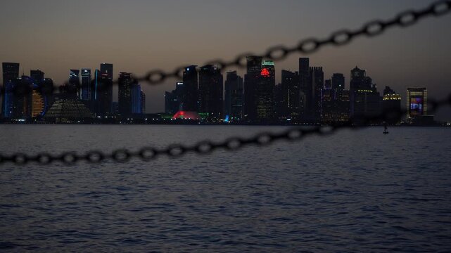 Cinematic view of the modern West Bay skyline in Doha, Qatar at dusk, creatively framed by out-of-focus nautical chains across the bay water.