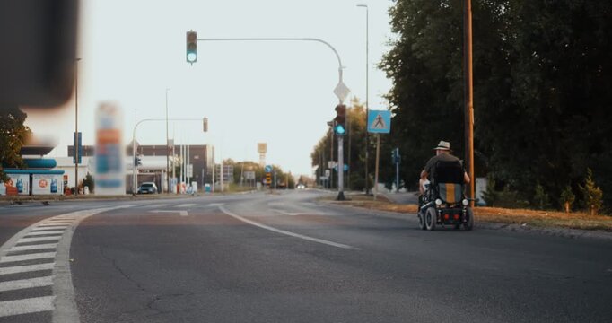 Rear view of a senior man in an electric wheelchair waiting at a red traffic light at a city intersection, showing independent urban mobility and everyday life.