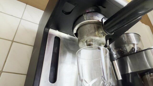 Low-angle close-up of a portafilter espresso machine brewing fresh coffee, with the liquid flowing from the machine before the brewing process stops. A coffee grinder is visible in the background.