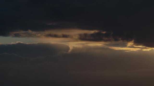 Narrow shafts of sunset light slip between dense storm layers, revealing shifting texture in the darkening sky.