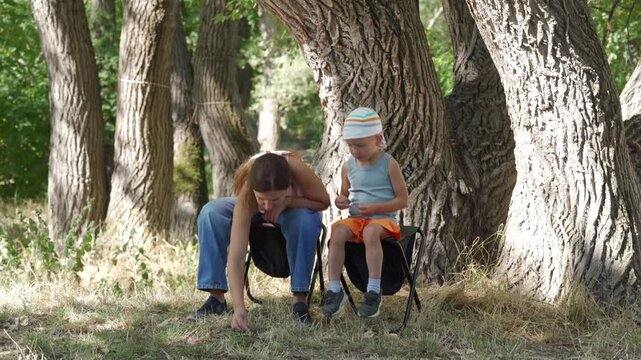 A tired mother and her calm son sit on folding stools under large trees. The mother reaches for the ground. They share a quiet, peaceful moment.