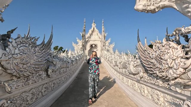 A blonde woman walks along a unique white bridge toward the entrance of a white Buddhist temple in northern Thailand, featuring ancient Thai cultural sculptures.