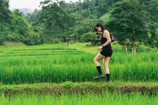 Young woman wearing black outfit and sunglasses walking carefully on edge of lush green rice field in rural outdoor nature setting