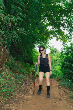 Woman hiking trail wearing black outfit using walking stick surrounded by green forest foliage on dirt path
