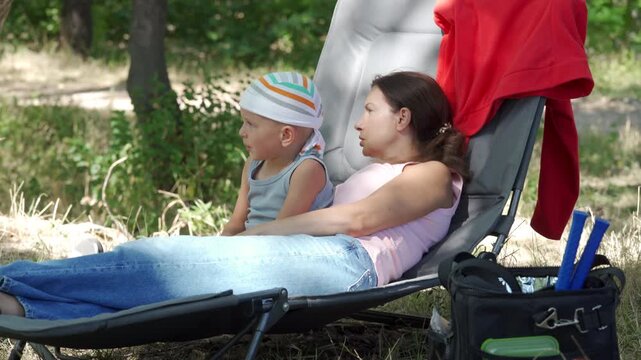Young woman resting on lounge chair beside child in shaded grassy park, with soft dappled sunlight, casual summer clothing, nearby towel, and calm family leisure in natural outdoor setting