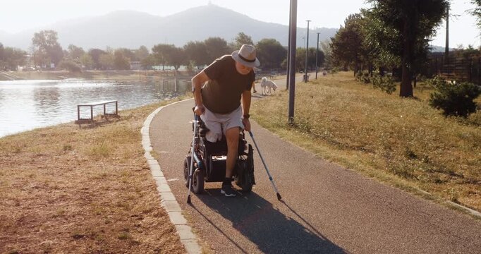 Senior amputee gets up from his electric wheelchair and walks with crutches beside a lake, practicing mobility and light exercise as part of an active daily routine.