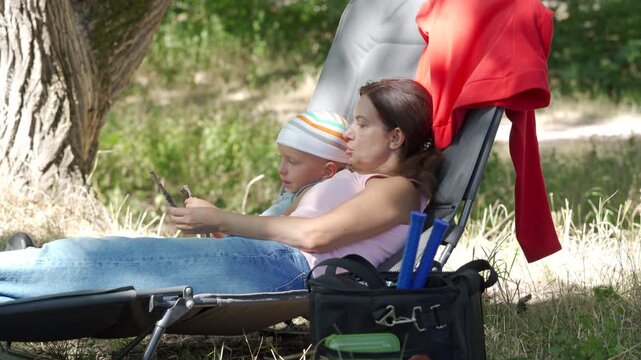 Young mother relaxes on portable lounge chair under tree shade while toddler rests close beside her, wearing sun hat in green park landscape with dappled sunlight and warm summer mood