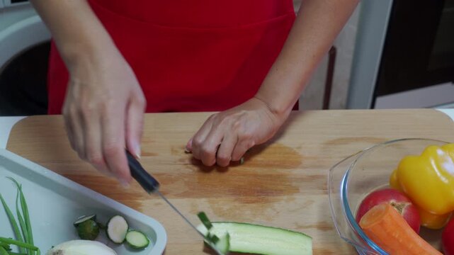 adult wearing red apron carefully chopping vegetables like zucchini, bell peppers, and carrots on a wooden cutting board and plastic tray in a kitchen setting viewed from an overhead angle.
