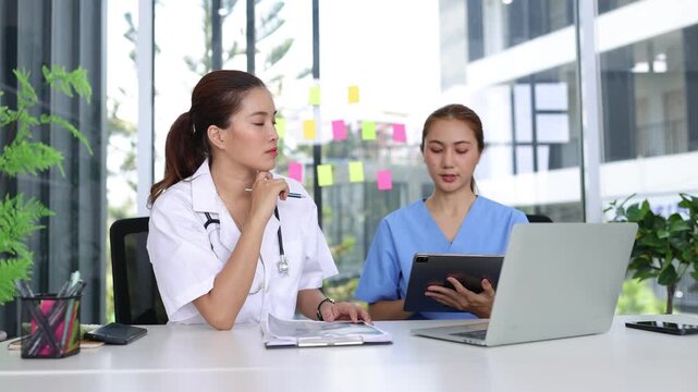 Asian female doctor and nurse discussing patient diagnosis with laptop and tablet in a modern hospital office.