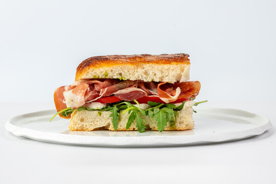 Gourmet Italian prosciutto sandwich filled with fresh mozzarella, ripe tomatoes, and arugula on crusty focaccia bread, served on a white plate in studio light.