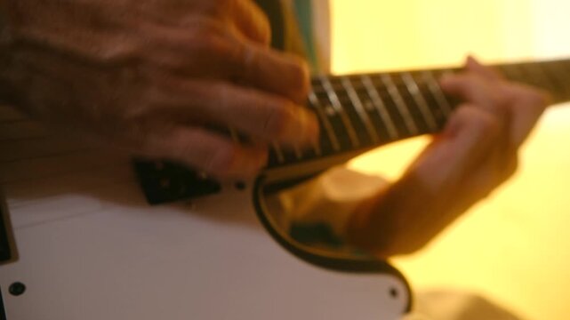 Close up man hands playing an electric guitar, fingers moving along fretboard and strumming strings during musical performance under warm yellow lighting in studio. Guitarist playing electric guitar