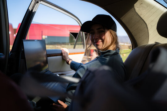 A woman is working on a laptop while traveling in a car, looking at the camera and representing the remote work, the digital nomad lifestyle, and work on the go.