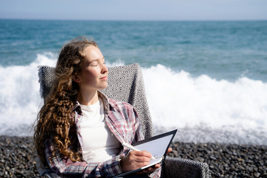 Woman journaling and sitting by the sea with a notebook, closing her eyes in a peaceful moment of reflection and enjoying the coastal atmosphere, wearing casual outfit.