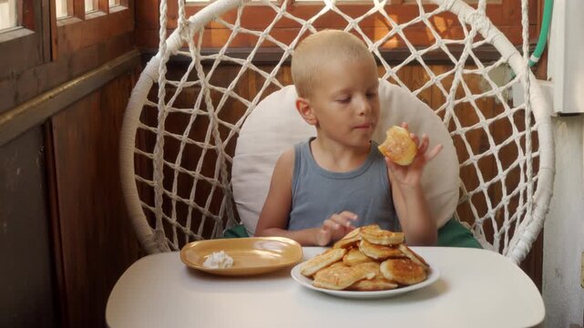 A happy boy eagerly eats a golden pancake at a white table. He feels joyful and content inside a cozy hanging chair. The wooden porch feels warm and peaceful.
