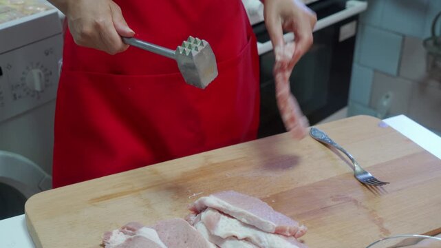 cook meticulously prepares meat in a kitchen setting, using a wooden chopping board and a metal meat tenderizer, wearing a red apron, with a focused expression, captured in a close-up view.