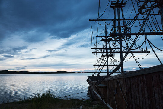 Blue-toned twilight lakeside landscape with antique-style wooden two-masted sailboats moored on the lake.