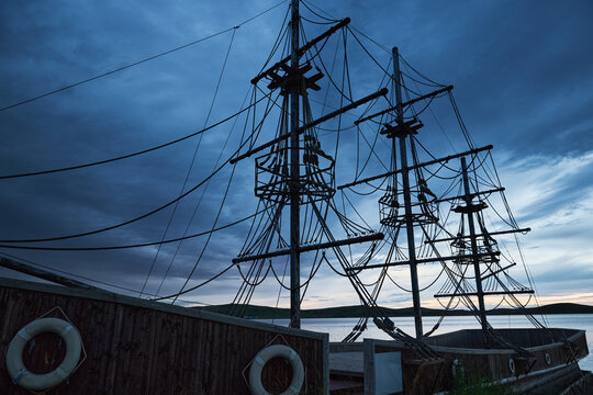 Blue-toned twilight lakeside landscape with antique-style wooden two-masted sailboats moored on the lake.