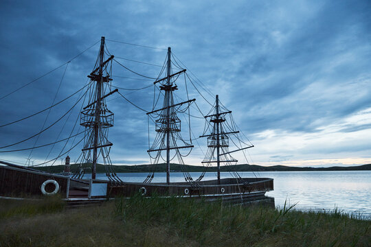 Blue-toned twilight lakeside landscape with antique-style wooden two-masted sailboats moored on the lake.
