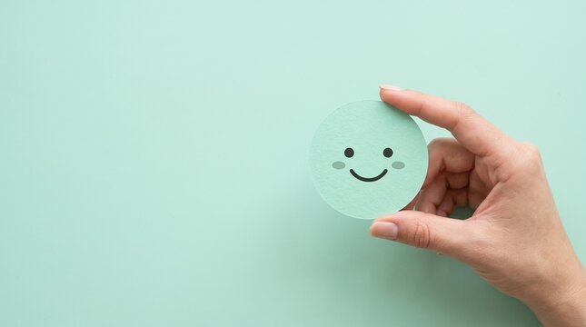 A human hand holding a cute paper cutout circle with a happy smiling face on a soft pastel mint green background for emotional wellness