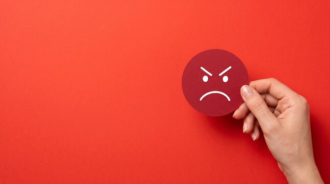 A female hand holding a paper cutout of an angry red face emoji against a vibrant crimson background representing emotional distress