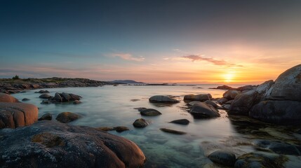 Obraz premium Majestic golden sunset illuminates calm coastal waters featuring smooth grey boulders scattered along the shoreline under a dramatic soft twilight sky during a peaceful summer evening