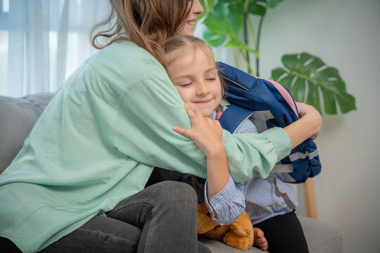 Emotional moment of mother hugging her little daughter wearing backpack on sofa. Warm embrace between loving young mother and cute elementary schoolgirl daughter in living room before leaving home.