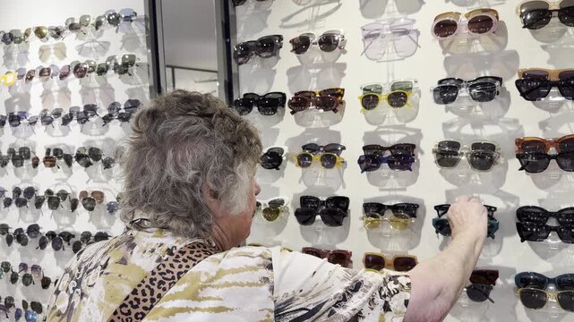 Senior woman browsing sunglasses at a retail display, looking at different styles and picking up a pair while shopping. Static shot.