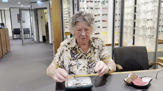 Senior woman seated at an optical store tries two different frames from an open case, putting aside the first pair before choosing and wearing sunglasses. Static shot.