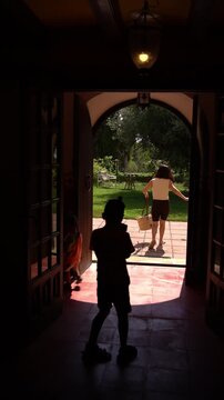 A mother walks outside into the garden while two children linger near the doorway, one following her and the other staying inside