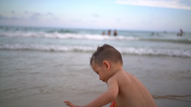 A toddler stands facing the sea, throws handfuls of wet sand and drops it into a bucket