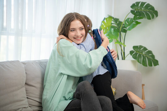 Cheerful young mom smiling while holding her schoolgirl daughter in warm embrace at home. Portrait of happy mother hugging her daughter with school bag sitting together on living room couch.