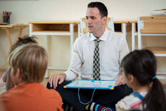 Male teacher holding small whiteboard teaching group of young elementary students in classroom. Male tutor holding whiteboard conducting interactive lesson sitting on floor with children in classroom.