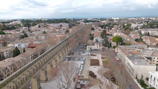 Saint Clement Aqueduct extends through Montpellier beside streets and urban buildings, aerial perpendicular approach establishing