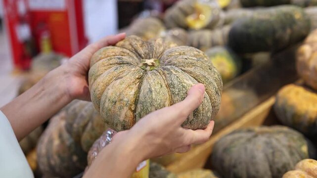 Close-up of a person holding and choosing a fresh ripe pumpkin or squash from a display at a supermarket or grocery store. Retail trade, food shopping and healthy nutrition concept.