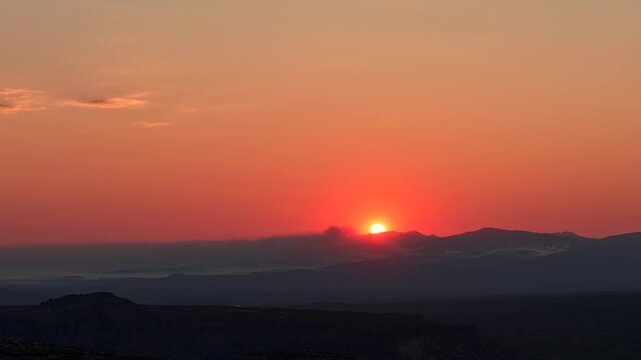 4K aerial timelapse of a blazing red sun cresting a dark mountain ridge. A thick, localized trail of wildfire smoke drifts across the peaks, contrasting sharply with the vivid orange dawn sky.