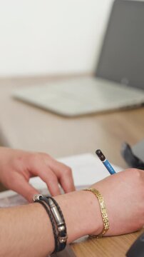 The framing highlights in the foreground a hand with leather and metal bracelets holding a blue pencil over printed sheets
