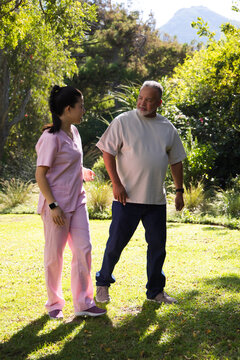 Asian nurse and African American senior walking and talking in park in pink scrubs and watches