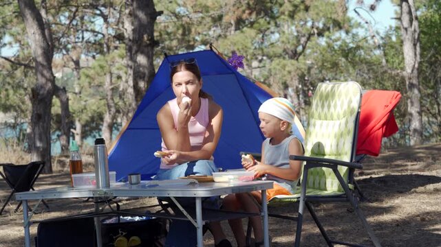 A relaxed mother and her content son happily eat a picnic lunch at a folding table. They feel peaceful bonding near their blue tent. The sunny pine forest campsite feels warm.