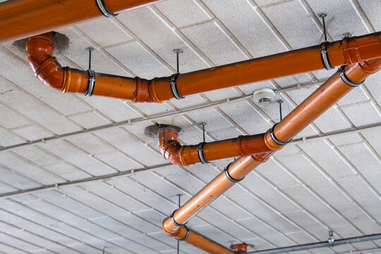 Orange plastic sewage drain pipes attached to textured concrete ceiling surface using metal clamps and hanger rods, featuring ceiling smoke detector in underground basement garage.