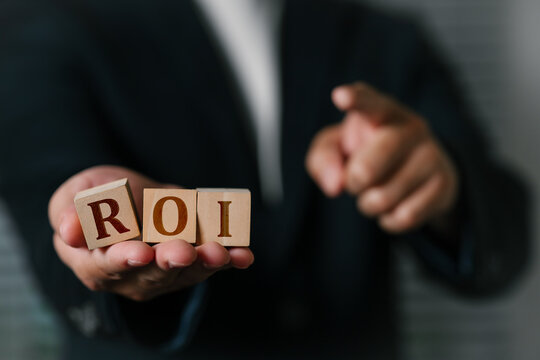 A business professional man dressed in formal attire holding wooden blocks spelling ROI Return on Investment, symbolizing financial growth, and success in the corporate world.