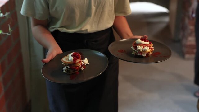 Person carries two black plates with stacked pancakes layered with cream and red berry sauce in a softly lit dining setting